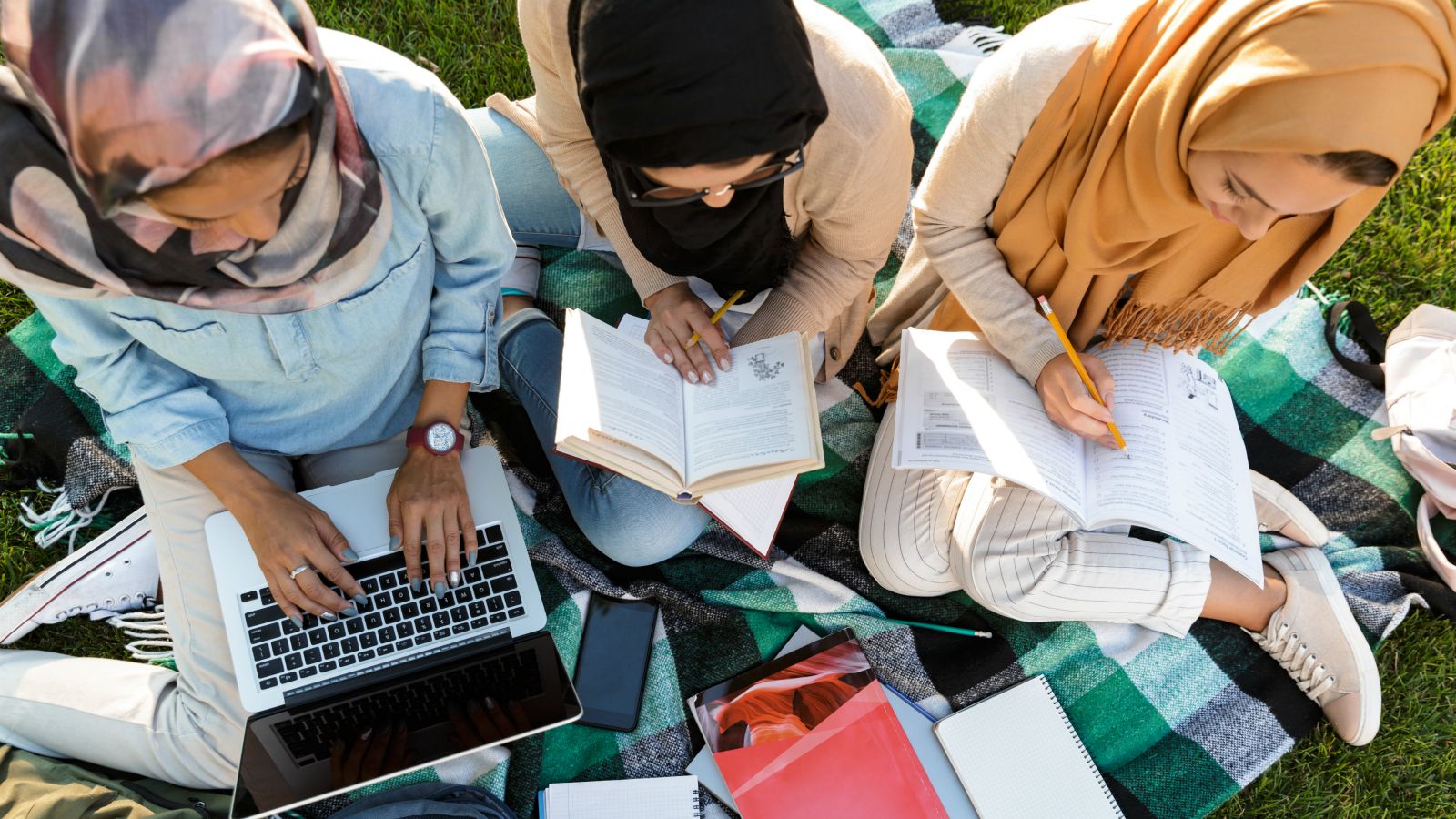 Saudi teachers sit outside on a blanket with a laptop, books and notes.