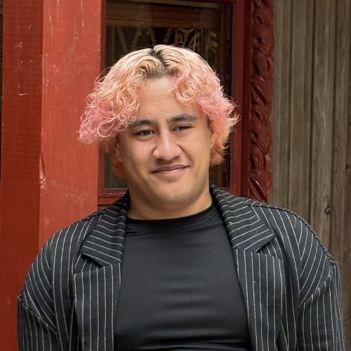 A person in a black t-shirt and blazer smiling, standing in front of a marae.