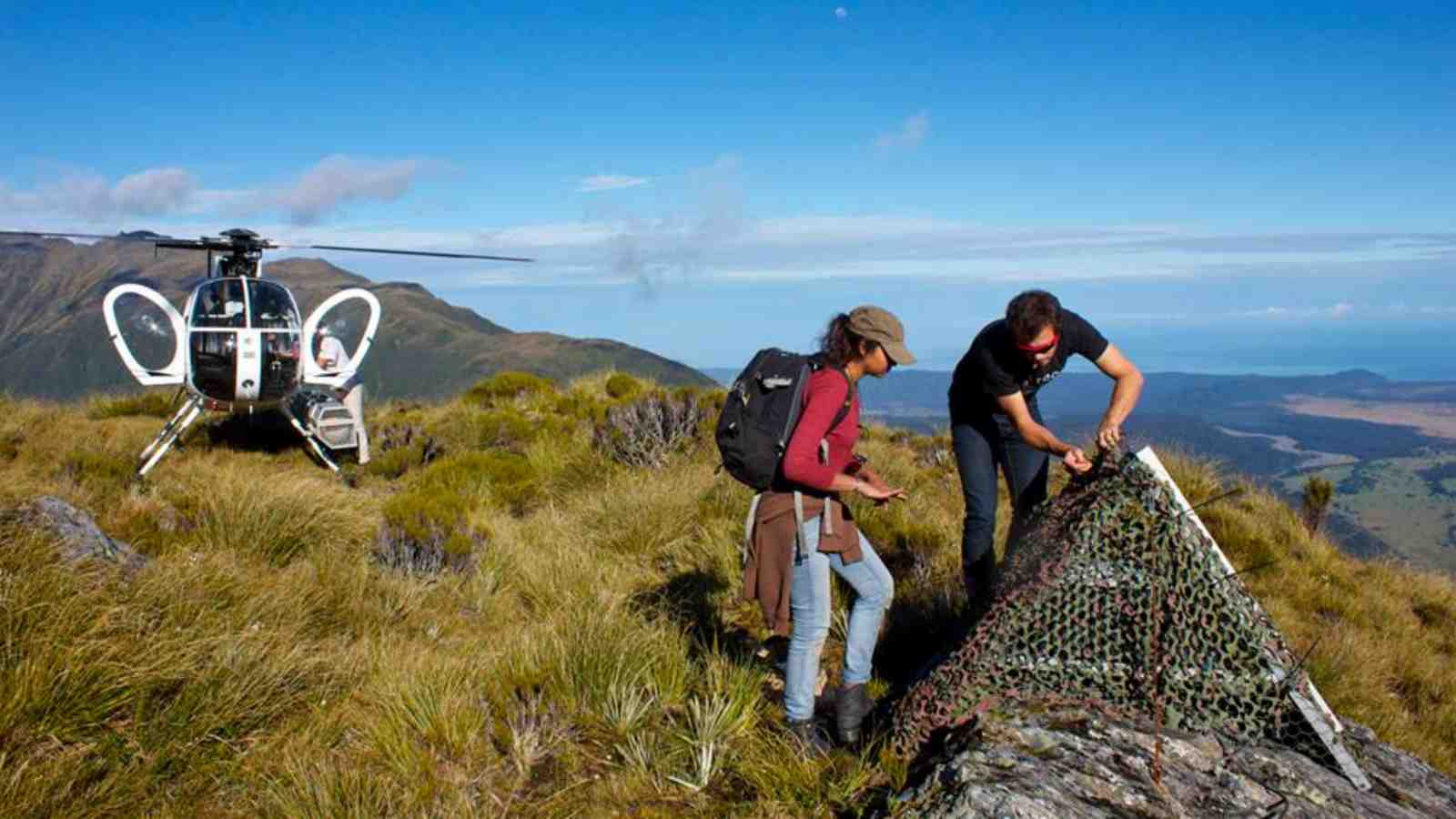 Dr Calum Chamberlain and PhD student Laura-May Baratin from Victoria University of Wellington look at monitoring equipment on Mt Baird in the Southern Alps. There are hills in the back and a helicopter near them. Photo: Dr Emily Warren-Smith.