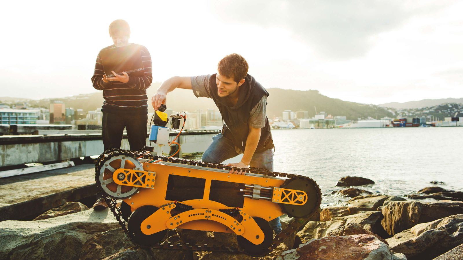 Two students work on an orange remotely operated vehicle that has large tracks – Wellington is in the background.