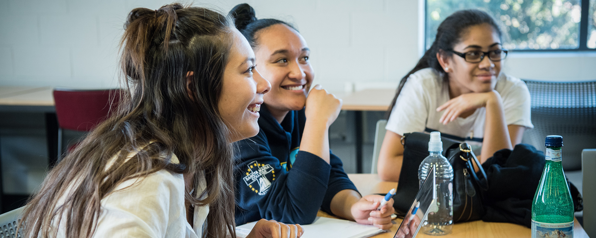 New students – three women smile together at a desk looking at a focal point out of frame.