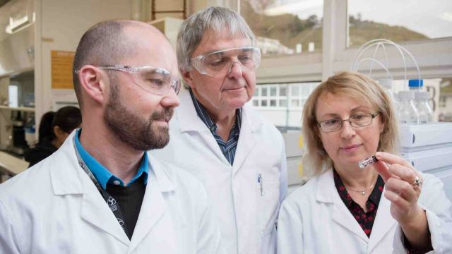 Scientists in lab coats inspect a small glass vile.