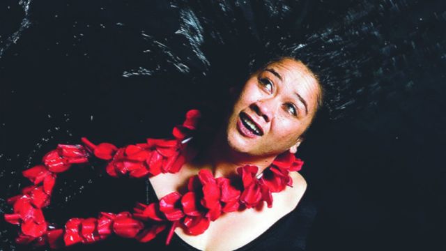 A woman wearing a red lei tosses her wet hair throwing water droplets that catch the light.