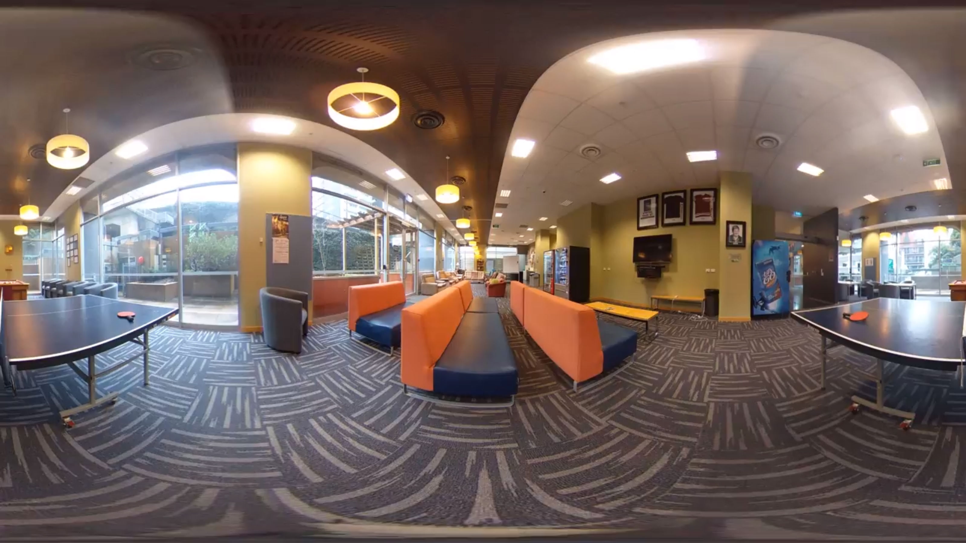 A panoramic view of a common space with orange sofas, a television, and ping pong table.