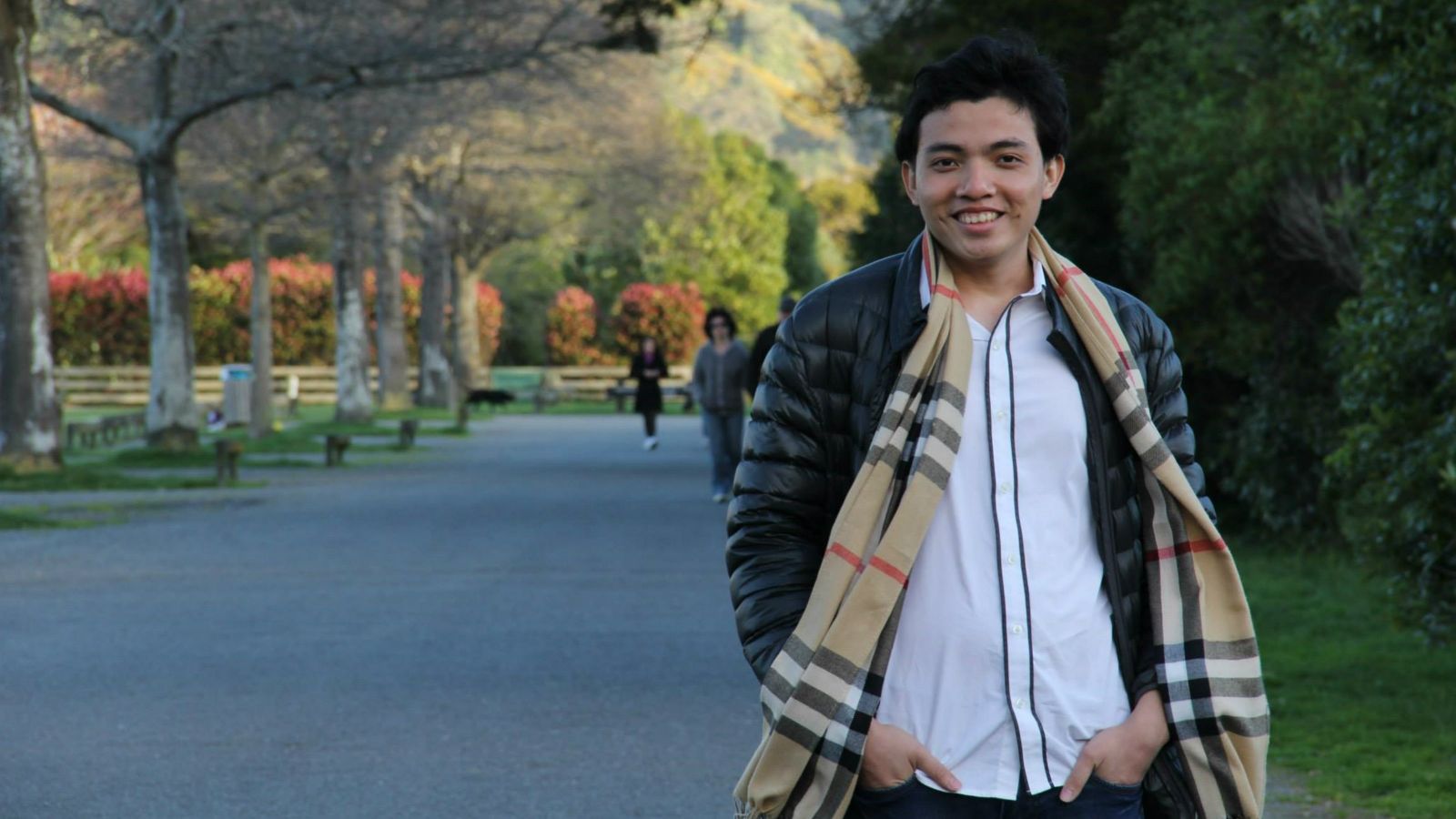 Wearing a white shirt, black jacket, and plaid scarf, Tran The Trung stands with his hands in his pockets on a park pathway shaded by autumnal coloured trees.