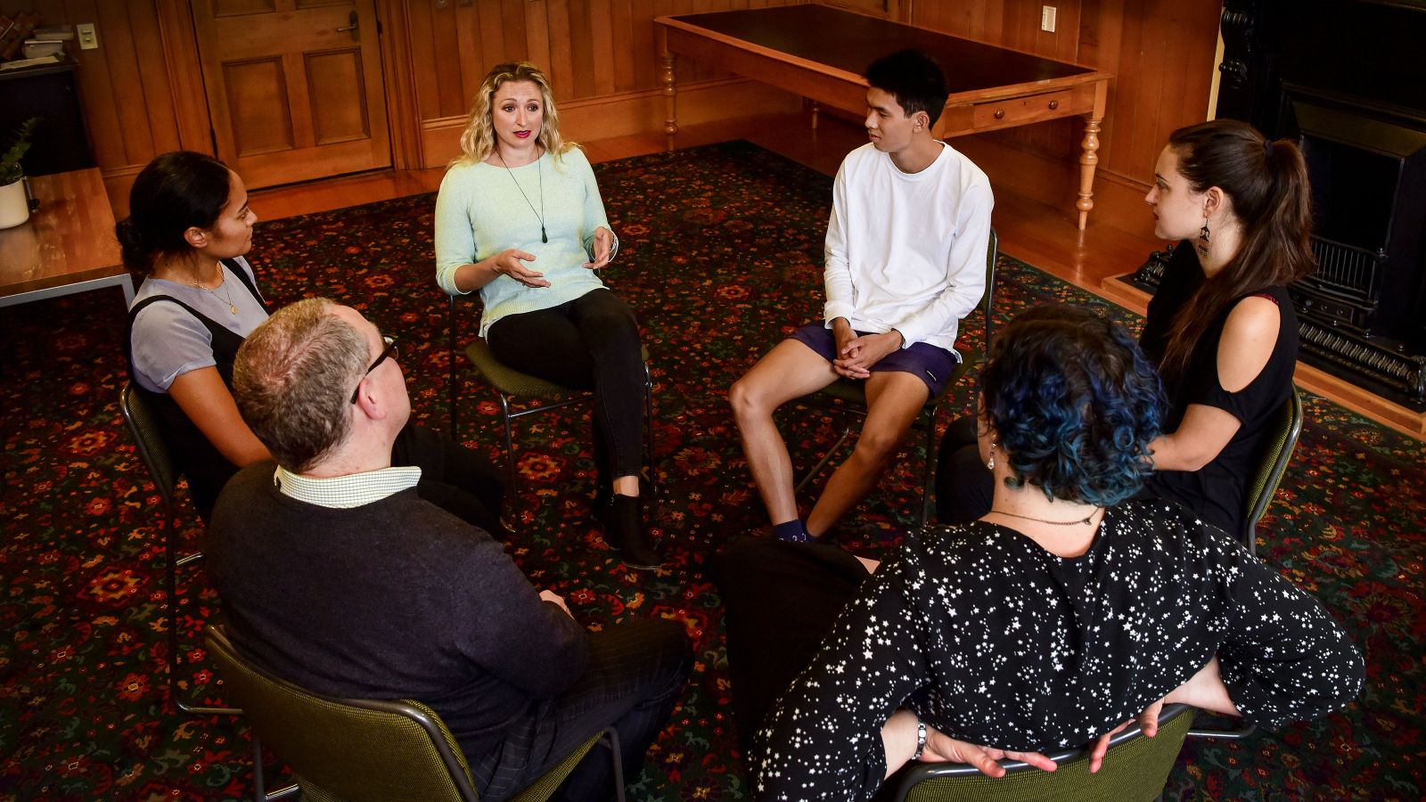 A group of six people, men, women, adults, and teenagers, dressed pretty casual, in shirts, cardigans, blouses, even shorts and sports shoes. They’re sitting in a circle in a red rugged, wood panelled room and having a discussion. Everyone’s attention is on one blonde women with an intent look on her face.