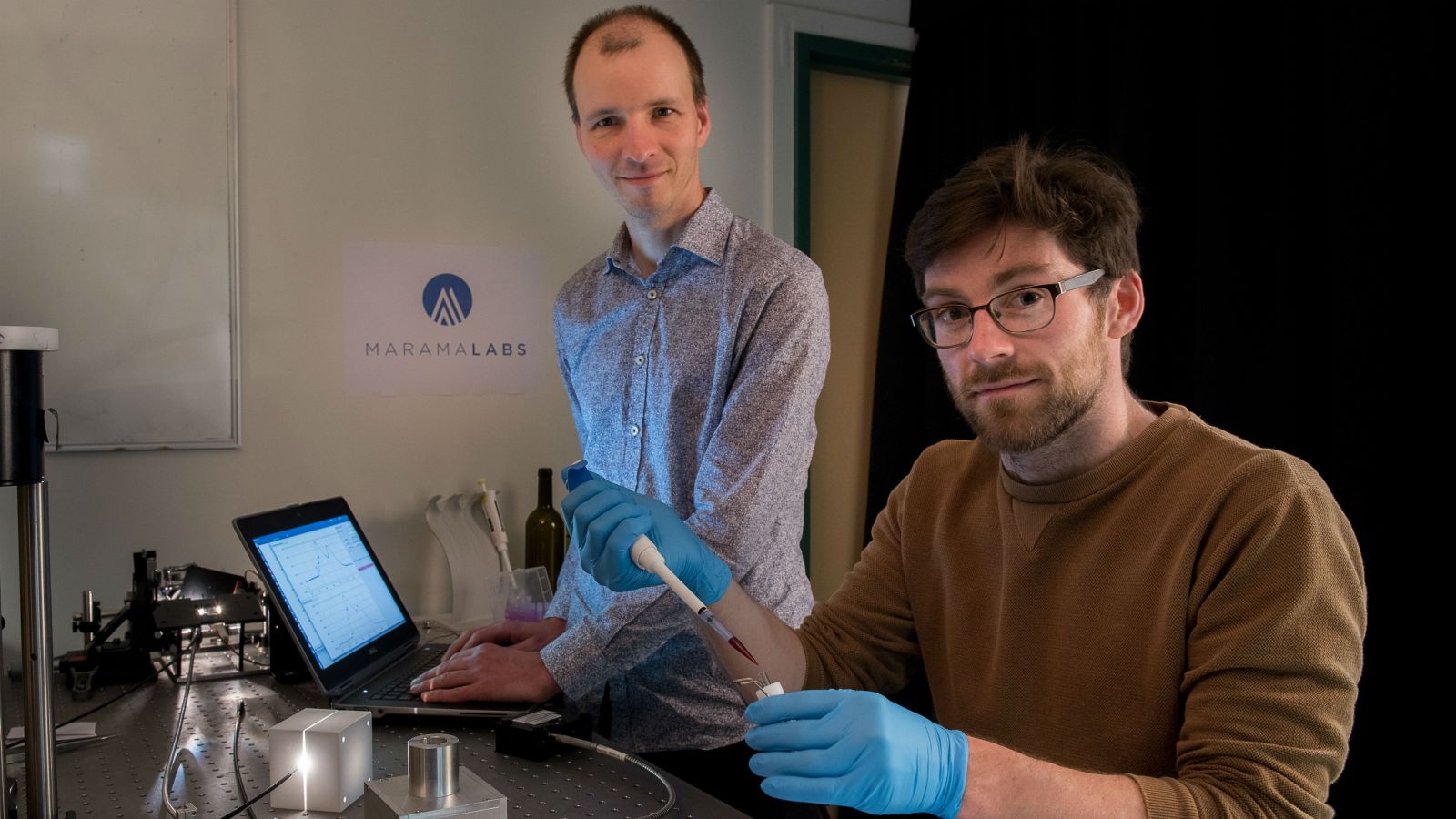 Mattias Meyer stands at a laptop with Brendan Darby sitting at the desk. Brendan wears blue gloves and is holding lab equipment. MaramaLabs and the logo are in the background. 