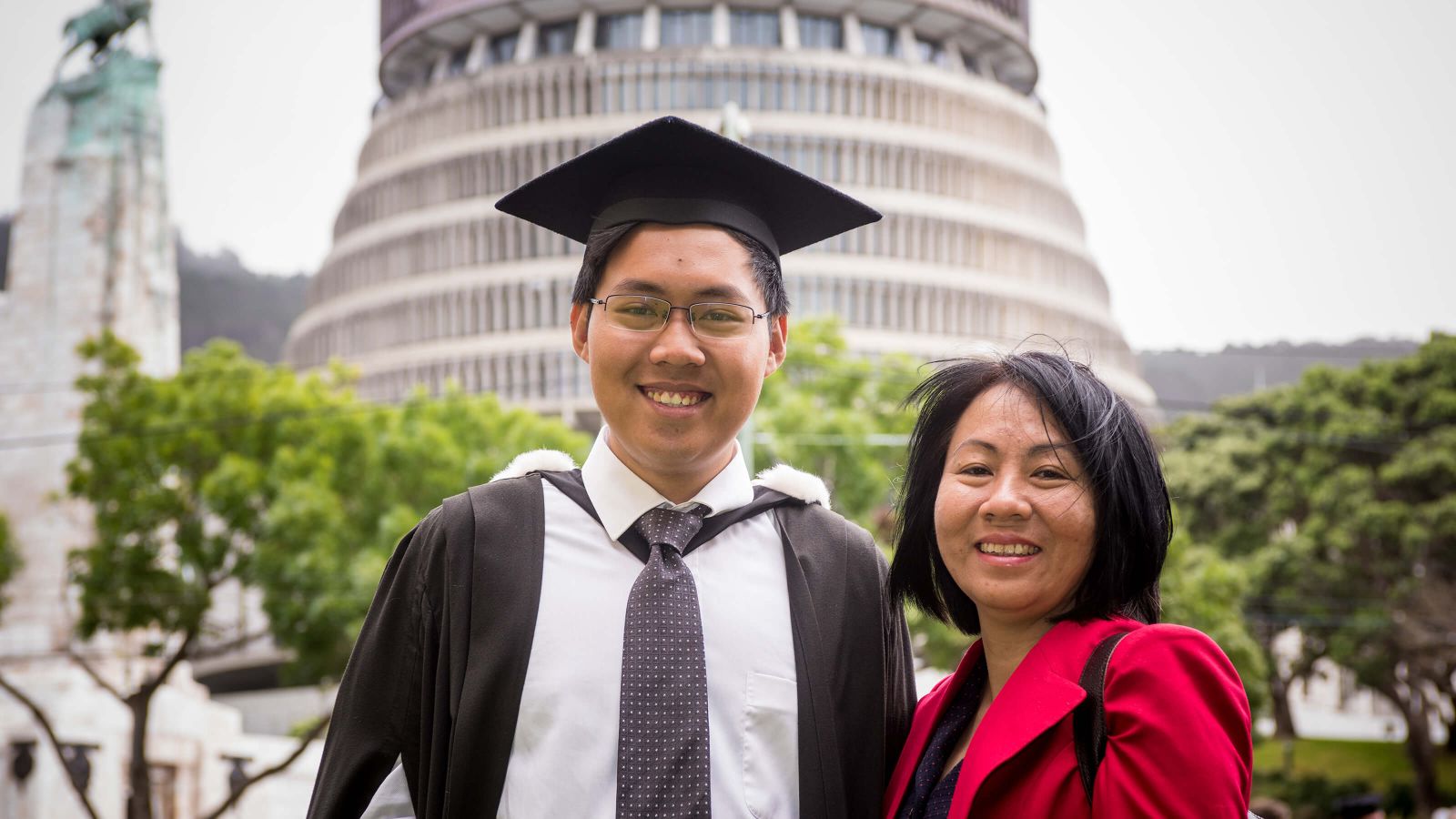 Male student with family member in graduation gown in front of parliament Beehive.