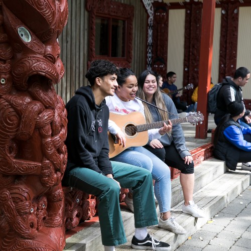 Two girls and a boy sitting outside a marae. One of the girls is playing an acoustic guitar and they are all singing.