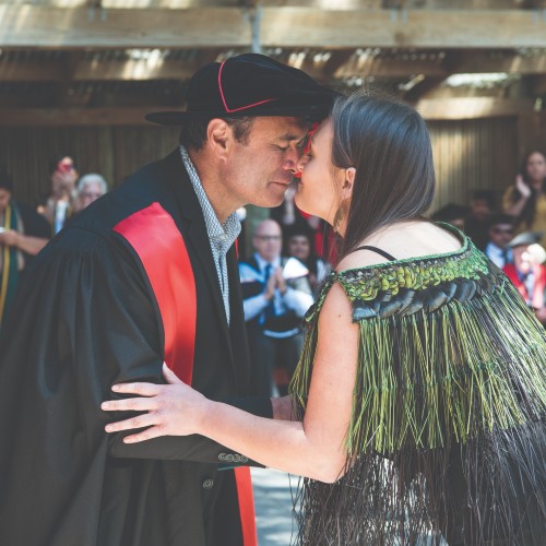 A man in graduation gown doing the hongi with a young woman wearing a Māori flax dress,
