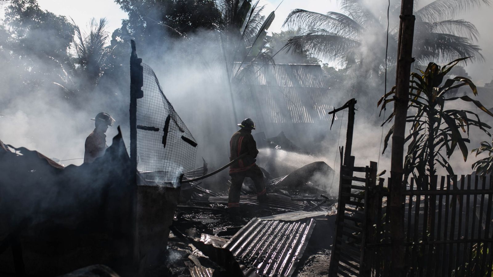 Silhouettes of two firefighters in helmets spraying water on burnt tropical shanty house with corrugated roof iron in foreground. Aftermath of fire in Philippines. Photo: Denniz Futalan, Pexels.