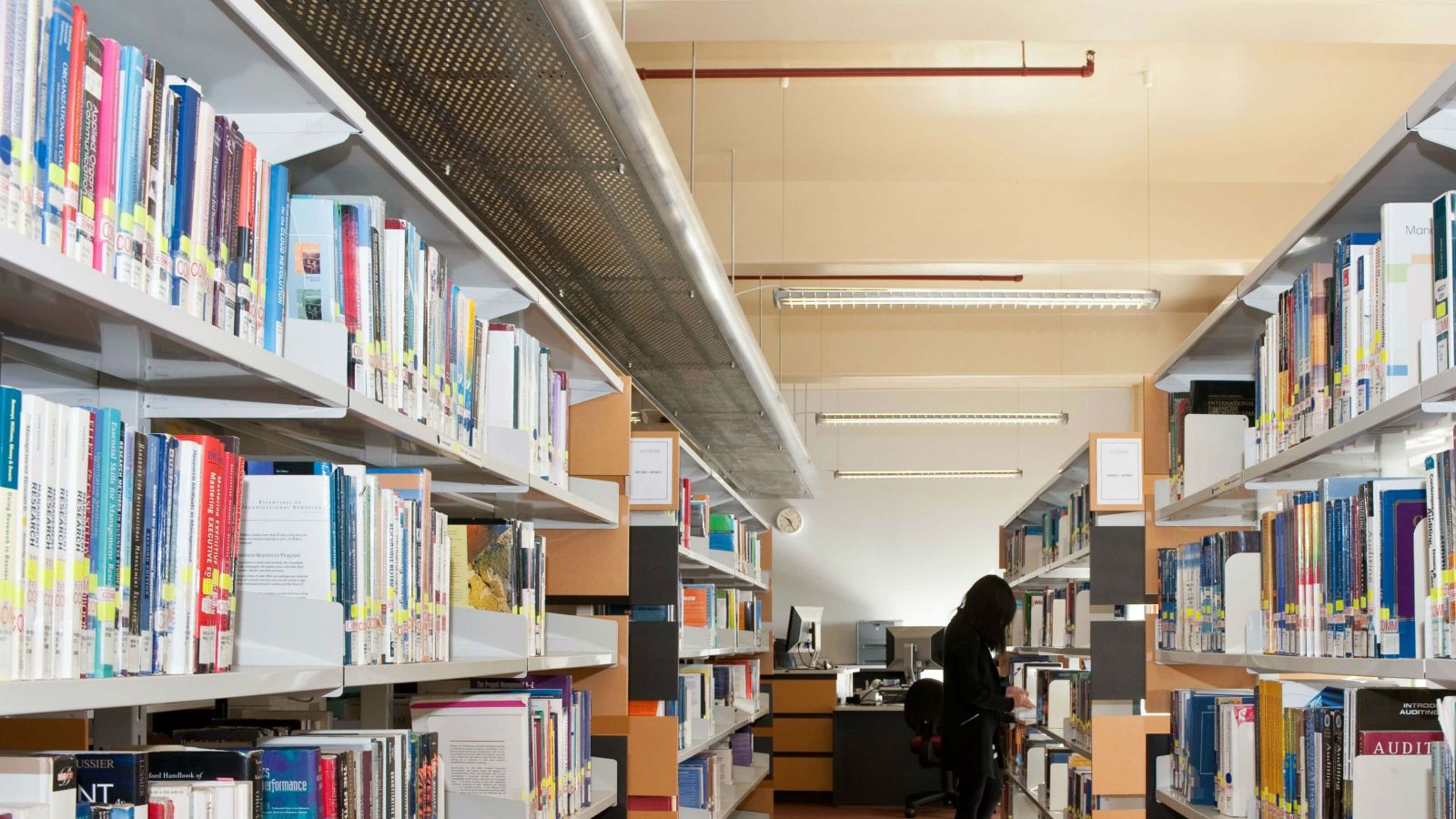 Books on shelves in a library. A student can be seen looking at books on one of the shelves.