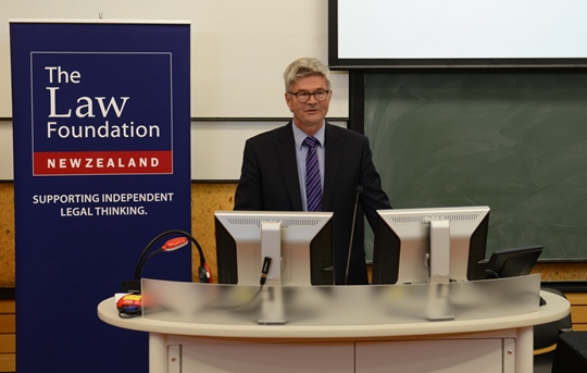A professional man speaks at the front of a lecture hall with the a poster in the background that reads, The Law Foundation of New Zealand, Supporting Independent Legal Thinking.