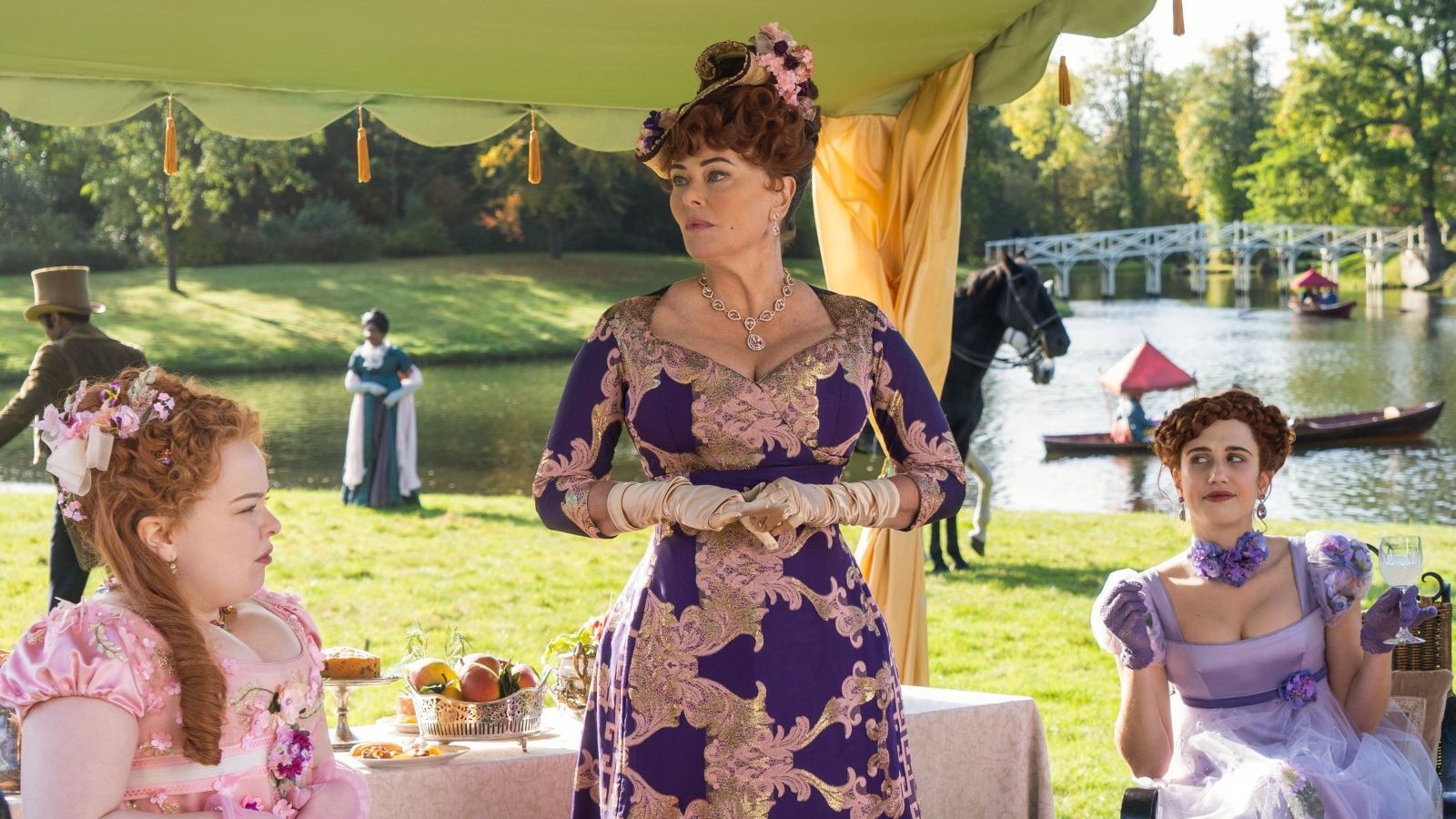 Three female Bridgerton characters relax underneath a gazebo by a lake