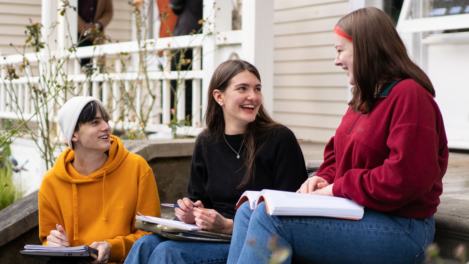 Three students with study materials sitting on the verandah of Helen Lowry Hall and talking to each other.