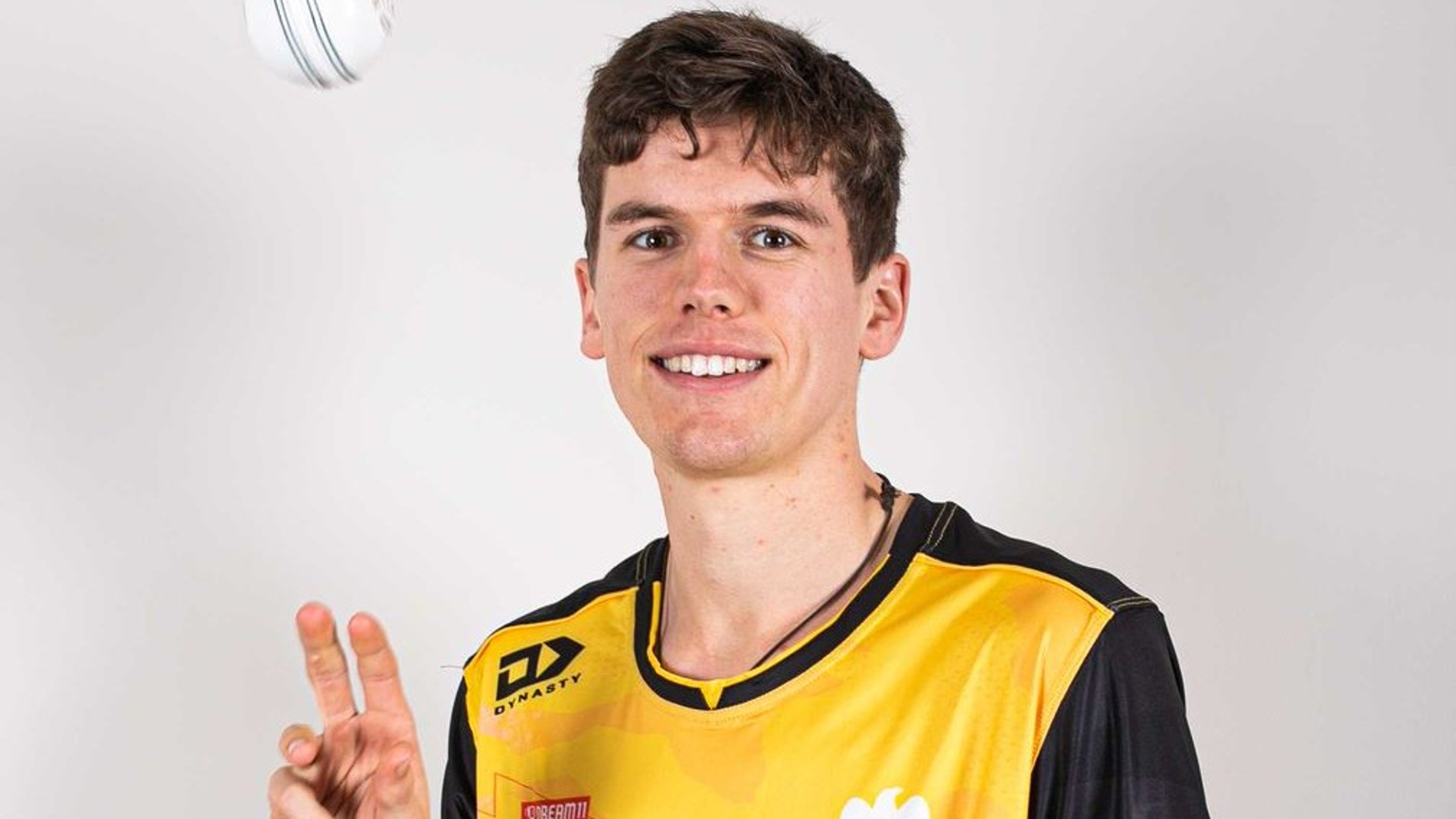 Young man in black and yellow sports wear tosses a white cricket ball