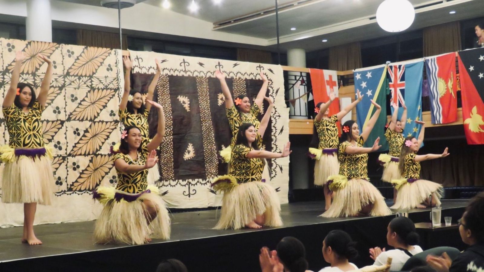 Dance performance on stage with tapa cloth in the background.