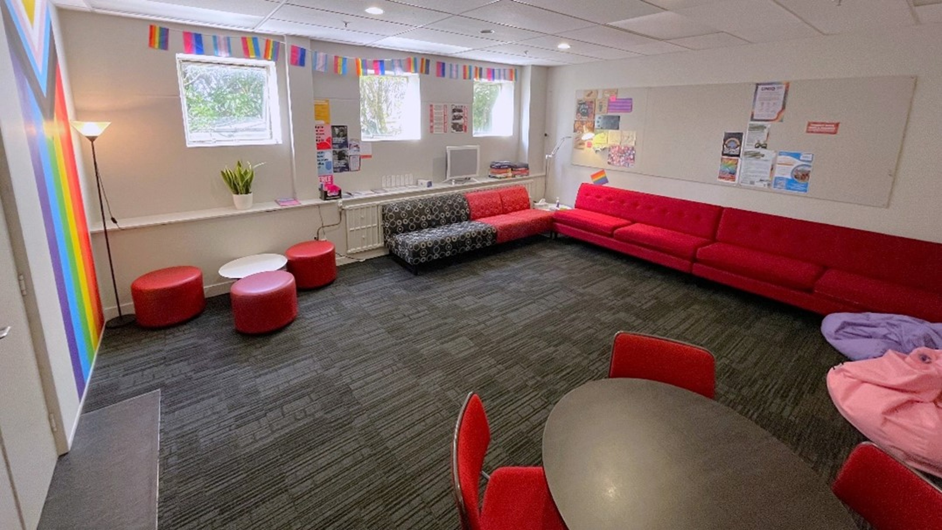 A well lit room with a dark textured carpet, red and black sofas, and a round table with red chairs. The room is decorated with LGBTQ+ pride flags.