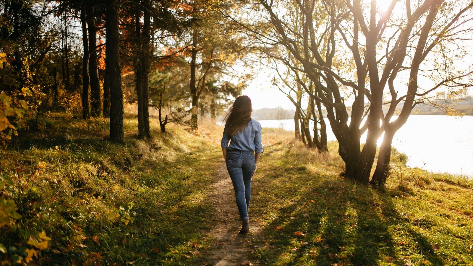 A woman dressed in blue denim jeans and a blue denim shirt, with long brown hair, walks along a forest path in autumn towards a lake, while walking away from the camera