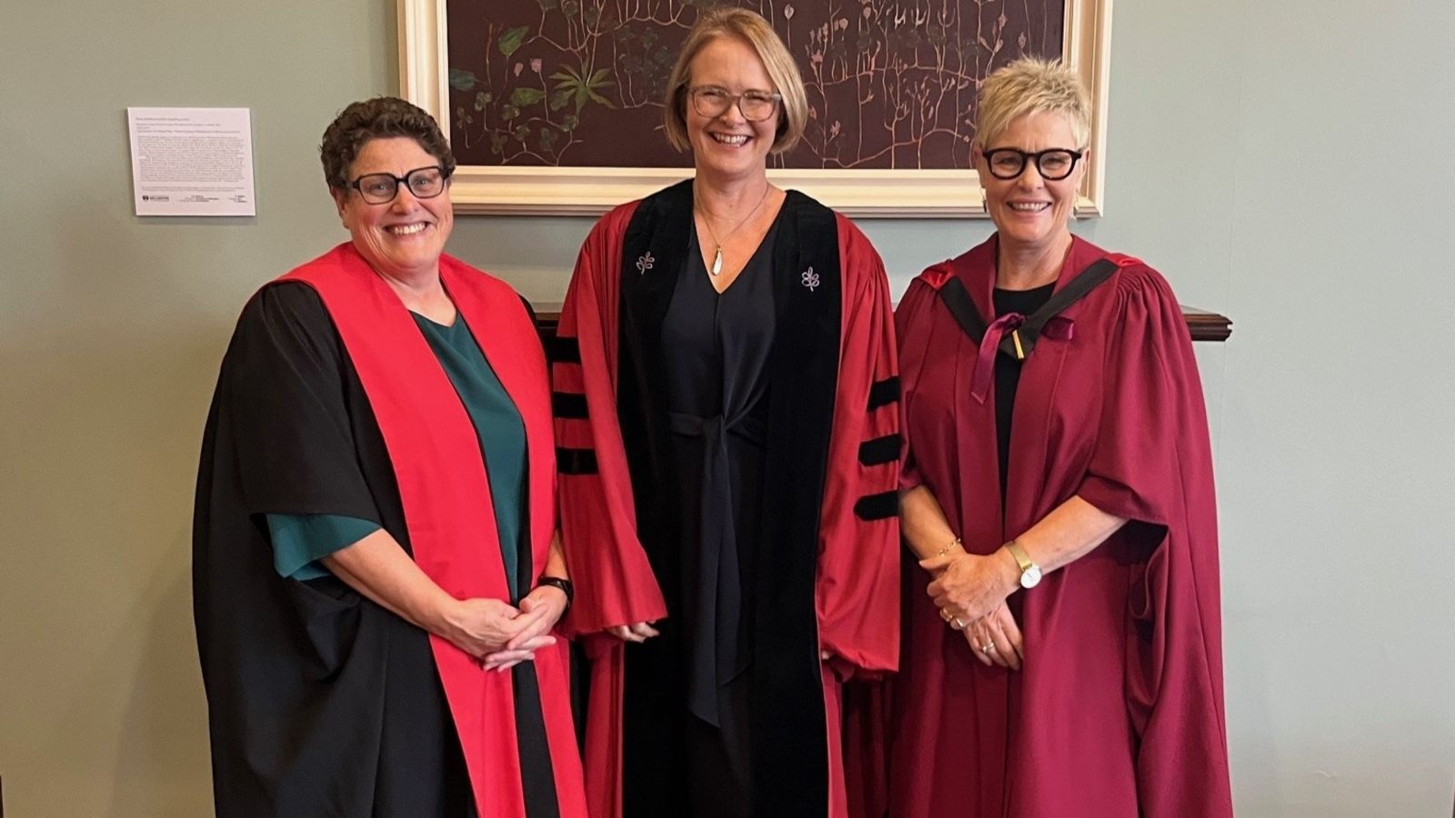 Three women wearing academic regalia stand smiling in front of a framed artwork on a pale green wall.