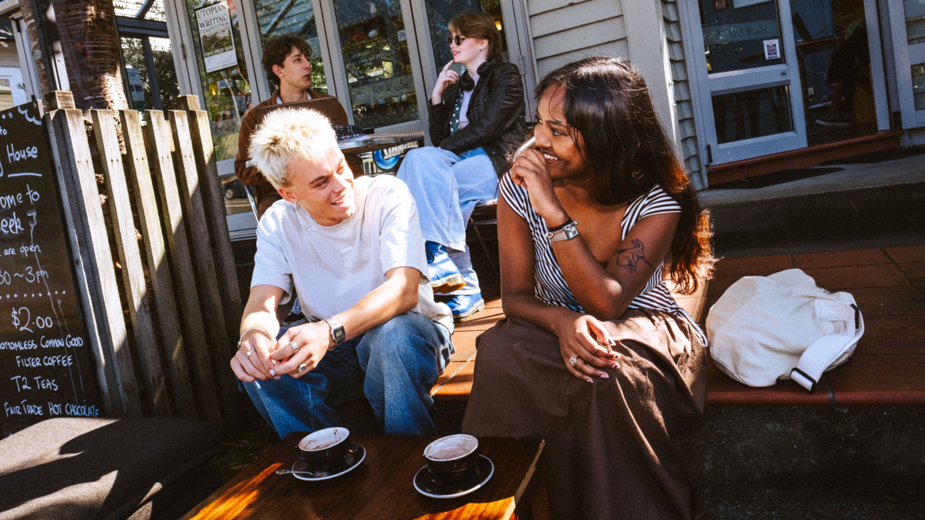 Two students sitting on the steps outside a campus cafe, enjoying a coffee and smiling at each other.