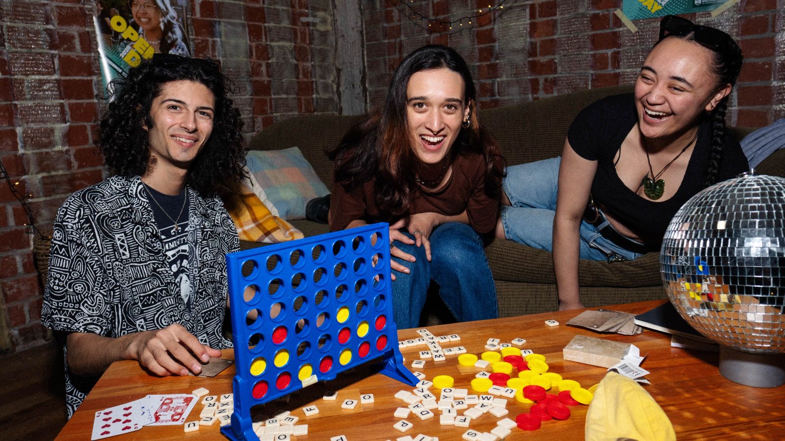 3 students sitting on a couch with connect 4, scrabble tiles and playing cards scattered on a coffee table