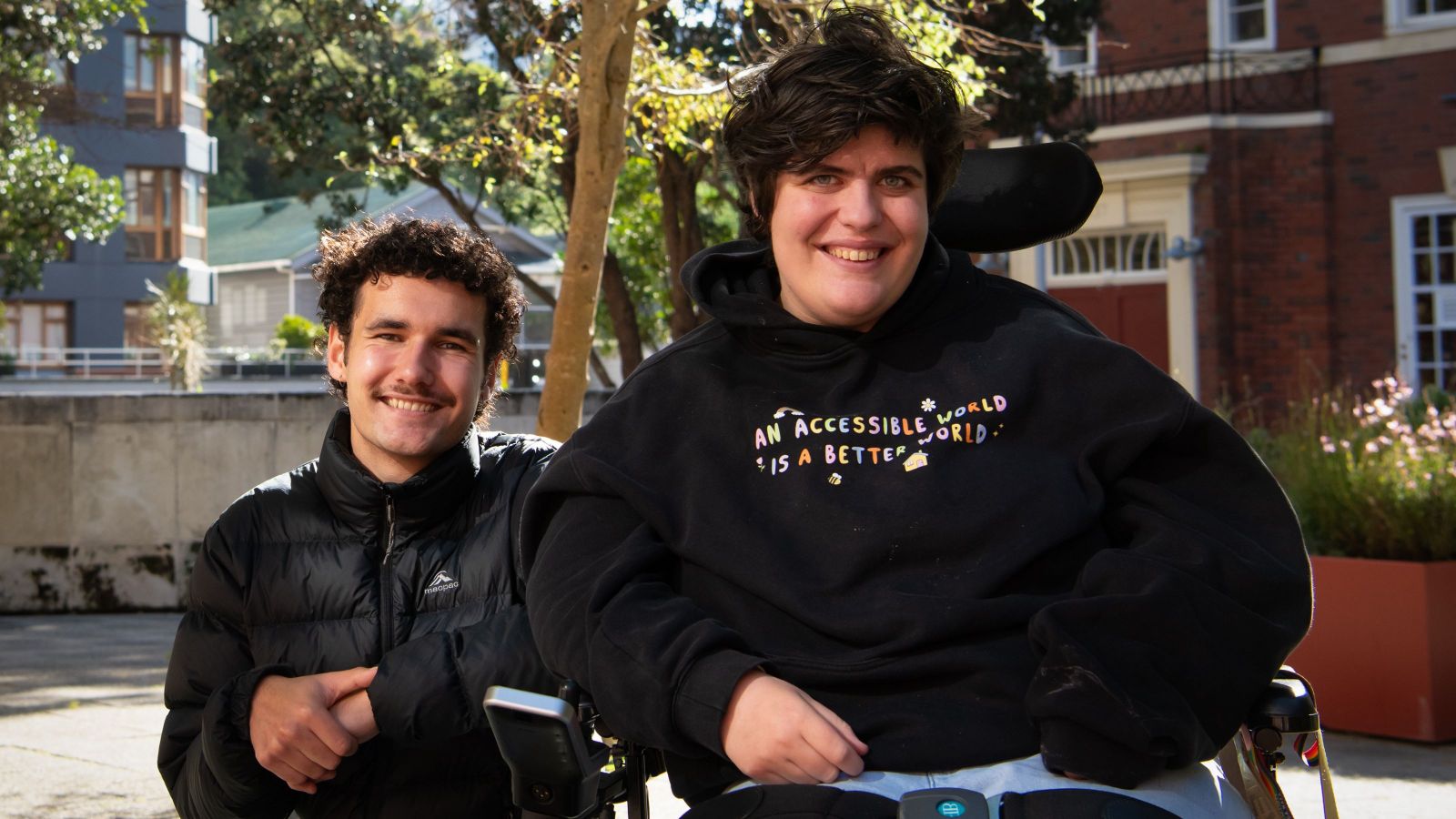 Josh and Etta, Rainbow Support Navigators, smiling together in the Hunter Courtyard.  Josh is kneeled next to Etta who is wearing a sweatshirt that reads 