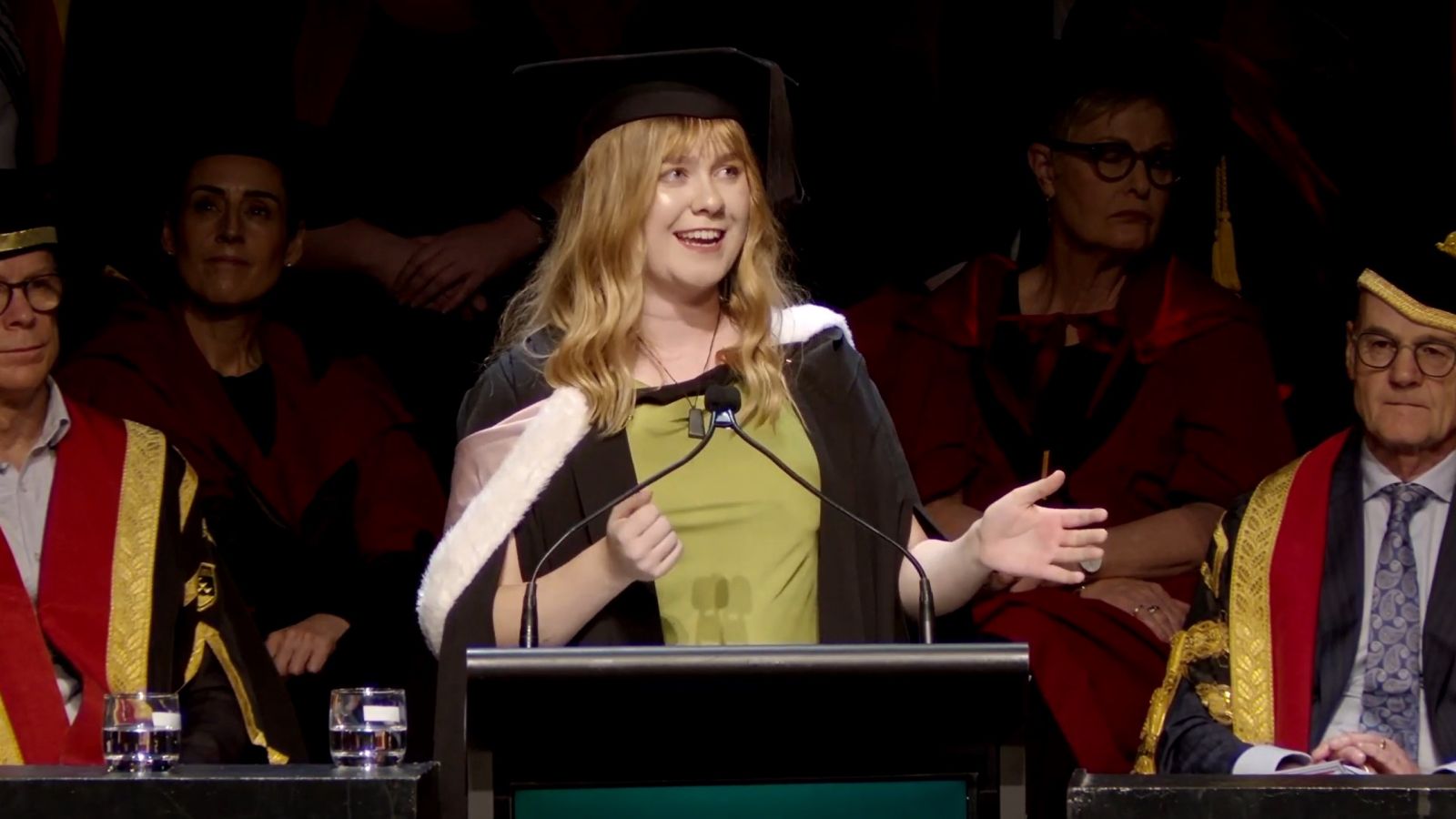 Graduate speaking at a podium during a graduation ceremony, with academic staff seated behind.