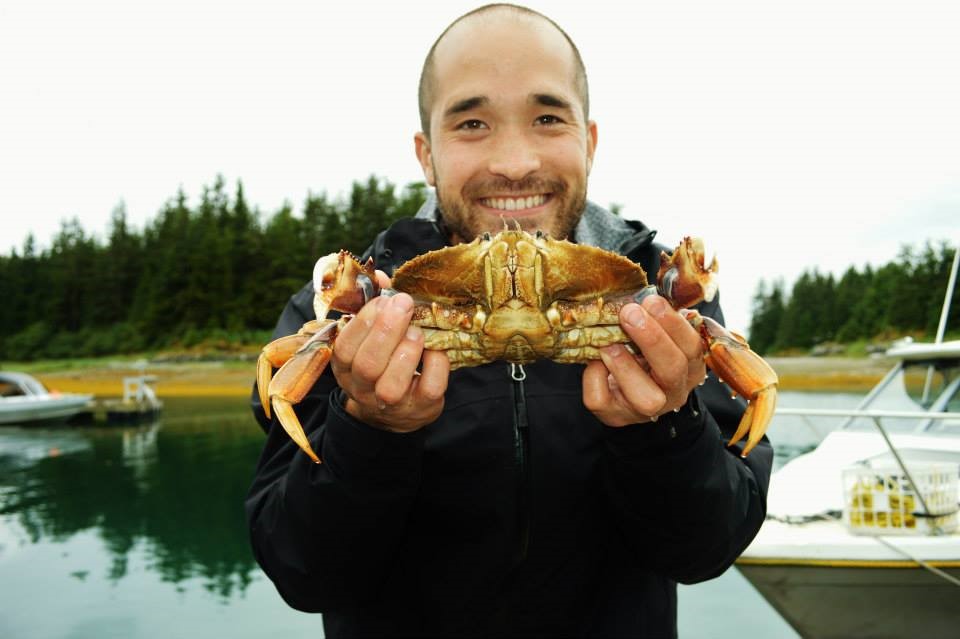 A picture of Jason Lescelius holding a crab while standing infront of docked fishing boats