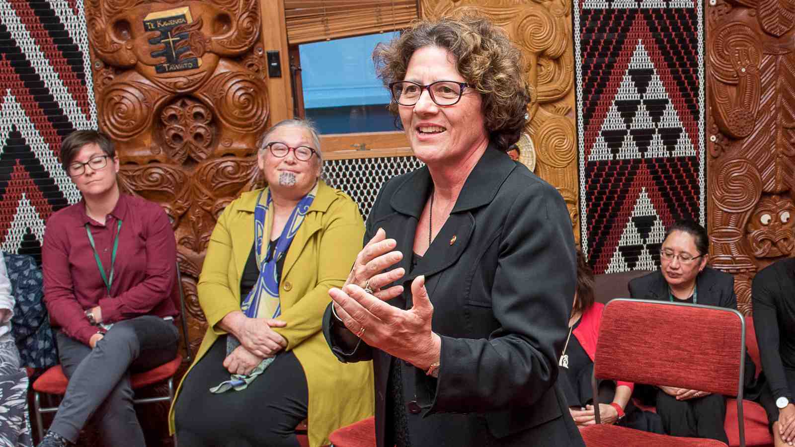 Bev Lawton stands in front of a crowd speaking at Te Herenga Waka Marae