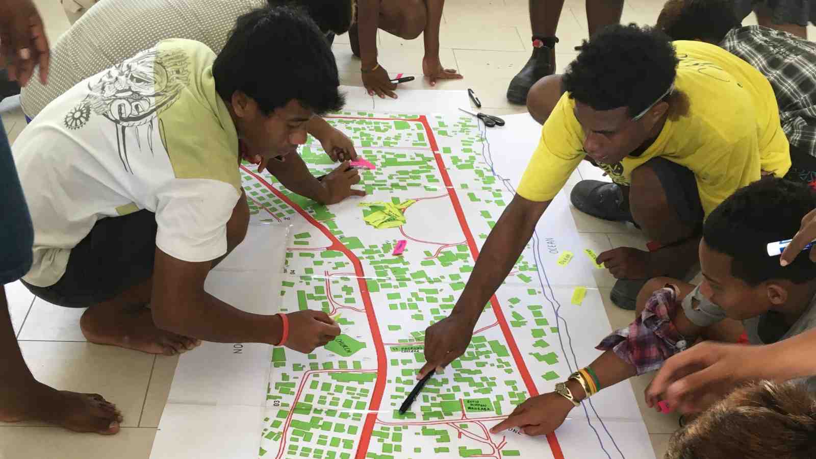 A group of youth from Betio, Kiribati sit on the floor around an architectural plan of their city, writing notes and planning their community centre. 