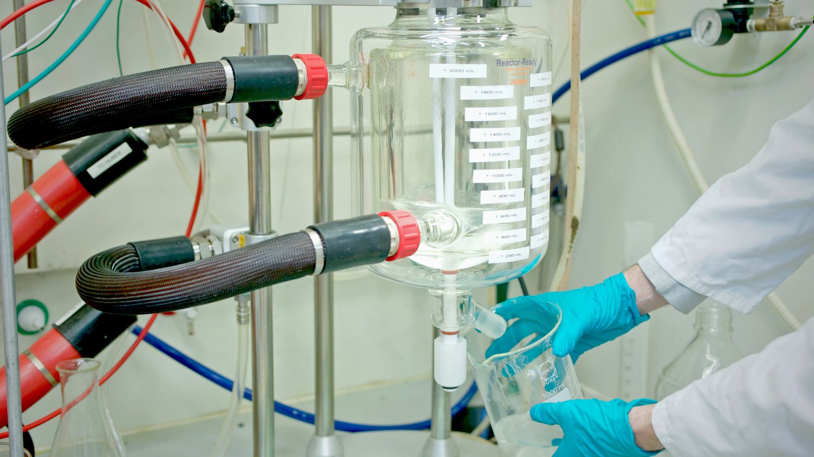 A scientist wears blue gloves and holds a beaker below a larger clear flask of fluid with hoses attached to it.