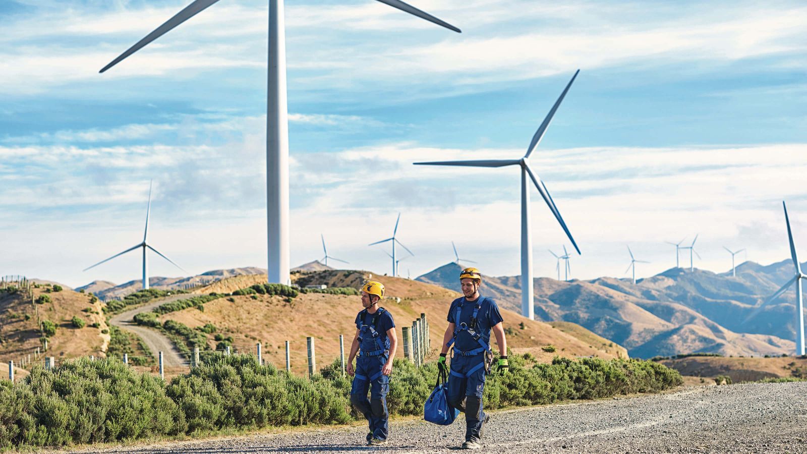 Two wind turbine workers wearing a safety harness and yellow hard hats, walk down a gravel road with many wind turbines in the background.