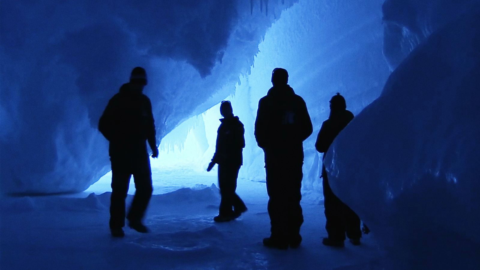Solid black human silhouettes explore an intensely blue ice cave.