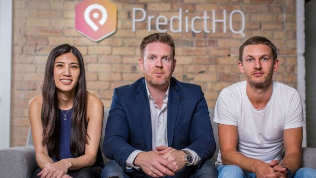 Yen Lim, Campbell Brown and Rob Kern sit next to each other on a sofa, with a brick wall, company logo and words 'PredictHQ' behind them. 
