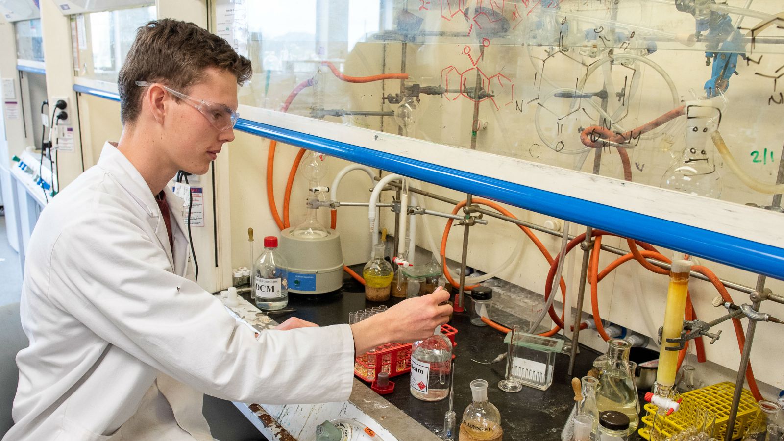 Matthew, wearing a lab coat and safety glasses, works with chemistry equipment. 