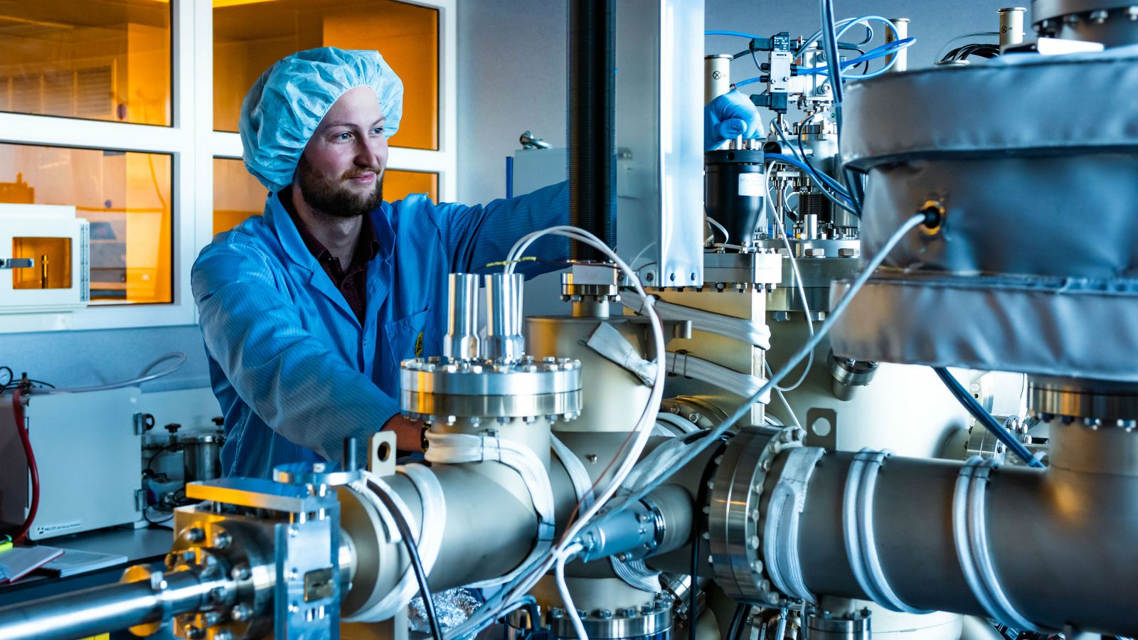 Tane, wearing a blue lab coat and a hair covering, looks at physics equipment in a small lab. 