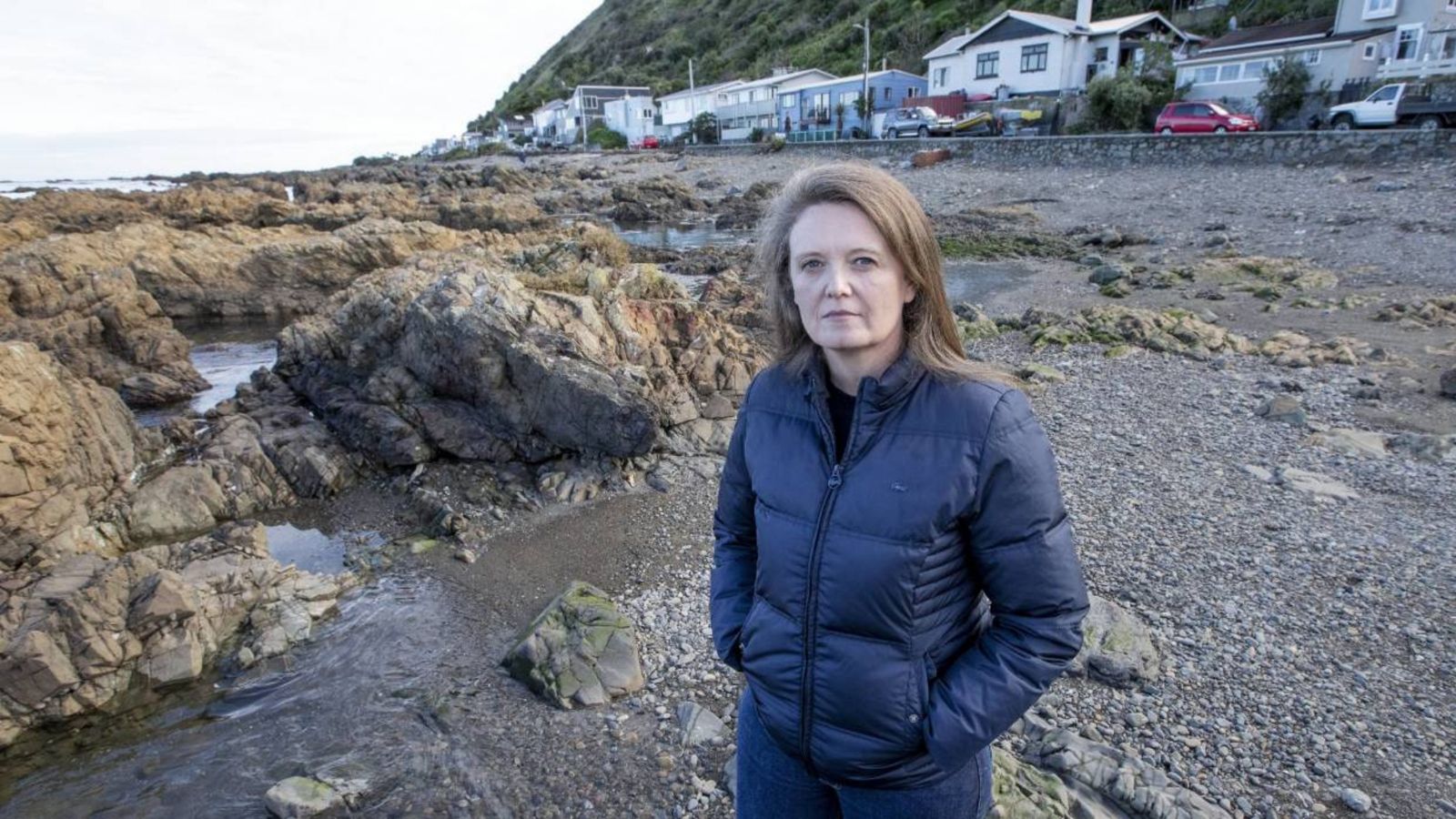 Phd candidate, Belinda Storey wearing a black jacket, standing on a beach in front of rocks on a cloudy day at Owhiro Bay. Houses and cars are on the street in the background. Photo: Monique Ford.