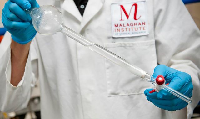 A scientist in a Malaghan lab coat works with a piece of glass laboratory equipment.