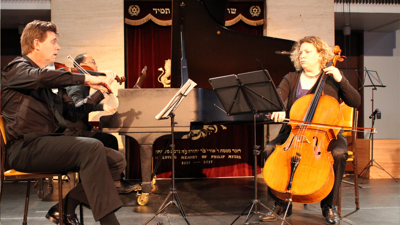 A man plays a violin and a woman plays a cello in the foreground with two music stands between them. Tucked in behind the violinist is a man playing a grand piano.
