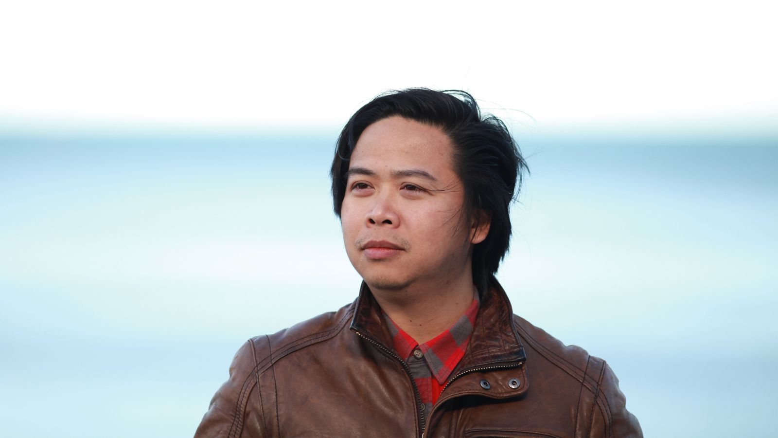 Quyet stands behind the ocean wearing a brown leather jacket looking away from the camera.