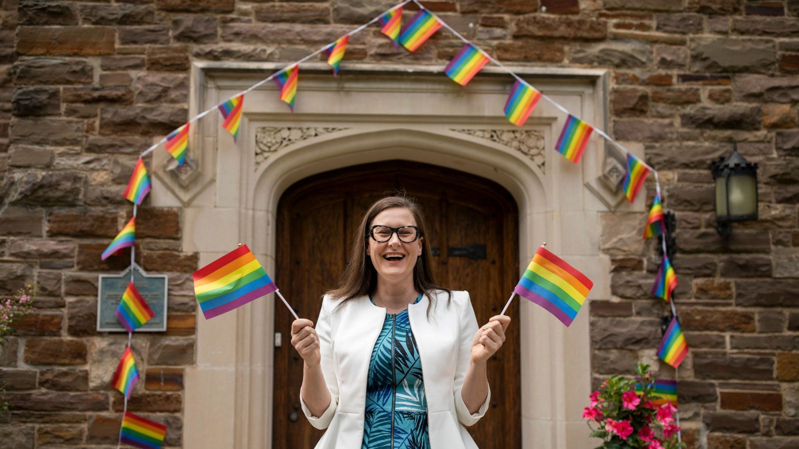 Woman with blue dress holding two pride flags, in front of old building covered in rainbow coloured flags