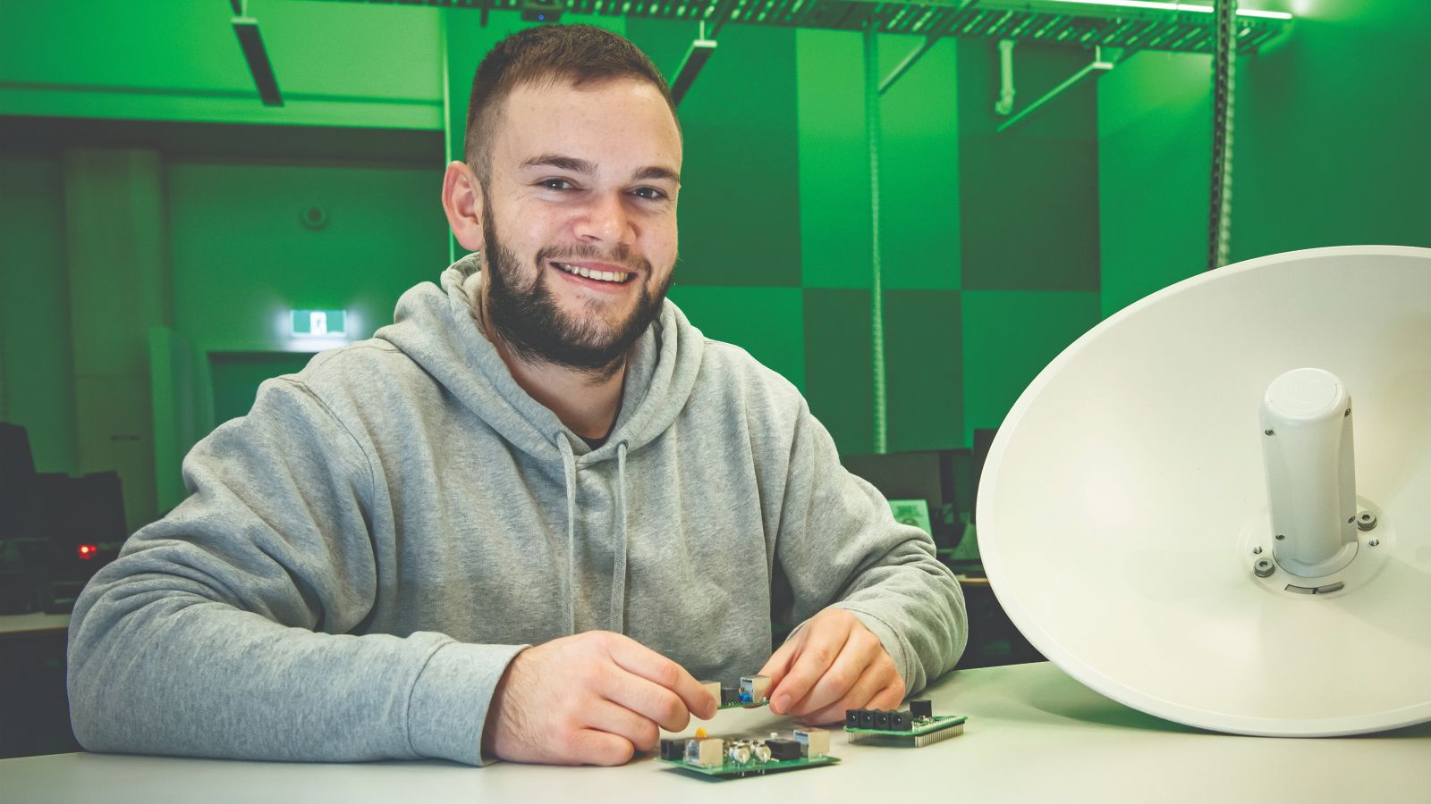 Duncan looks into the camera. He is holding a circuit board. The room is lit up in a green hue.