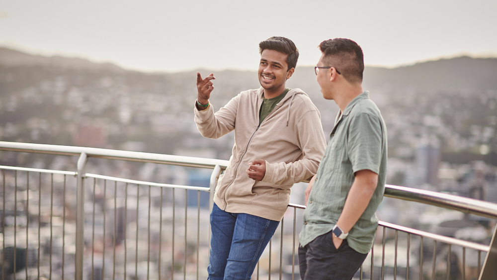 Two students stand smiling and talking with the city in the background.