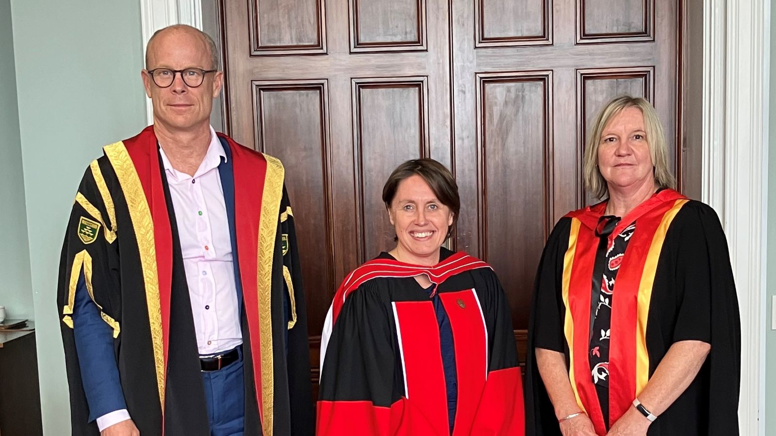  Nikki Hessell, flanked by Nic Smith and Sarah Leggott. All three are wearing academic robes and standing in front of a large wooden door.