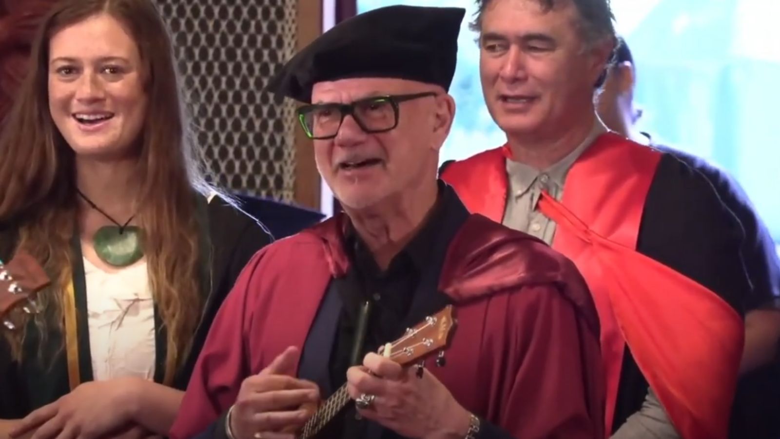 A graduate in red regalia plays the ukulele while university staff members in the background sing along.