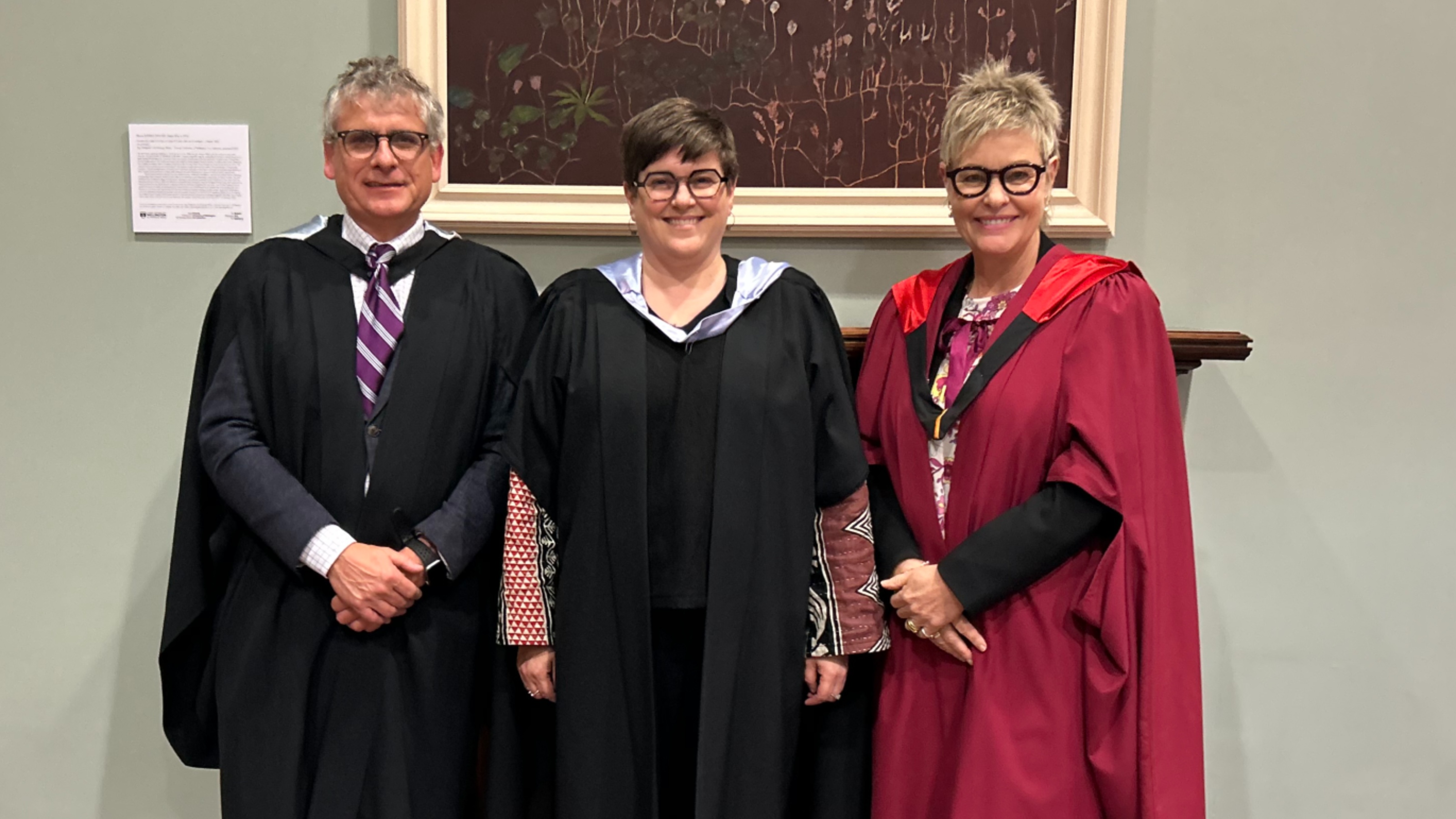 Three academics, one male, two female wearing academic dress in front of a pale green wall