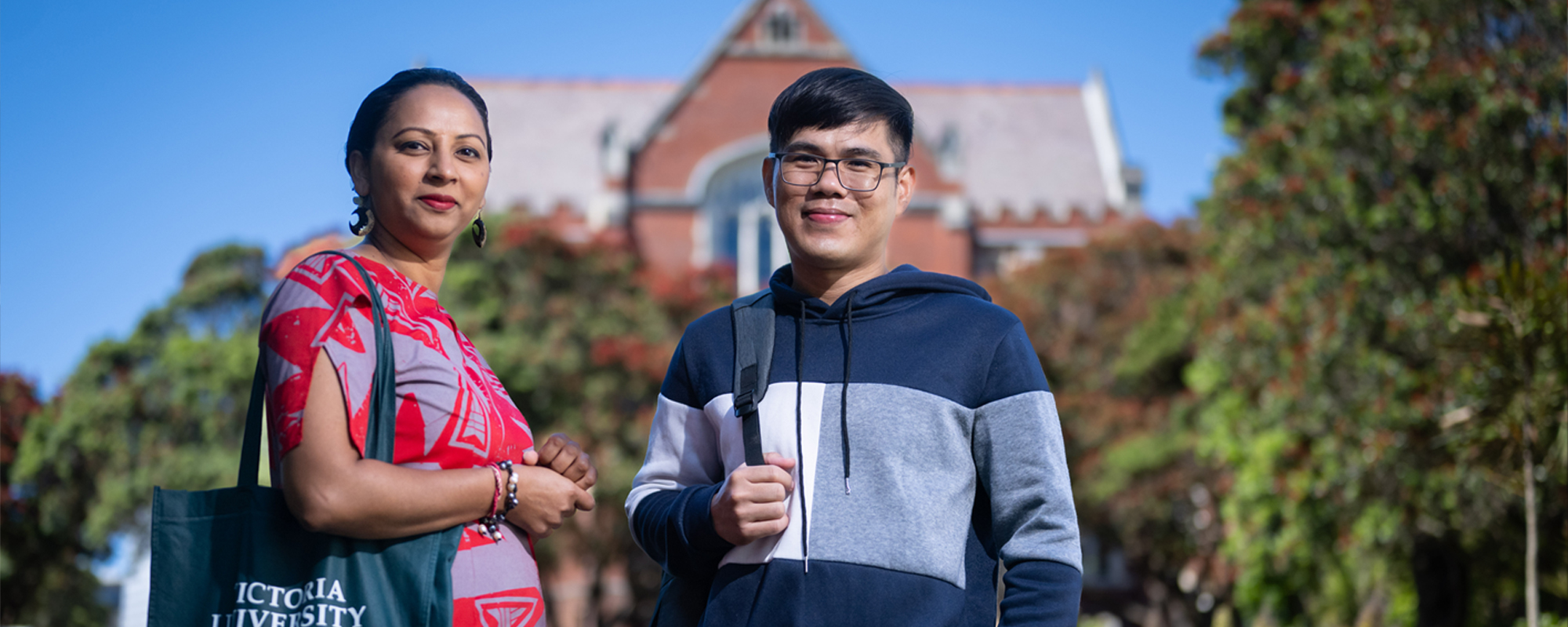 Two students from the Manaaki scholarships programme standing in front of the Hunter Building, which is a historic red brick building part of Victoria University of Wellington, in the Kelburn Campus.