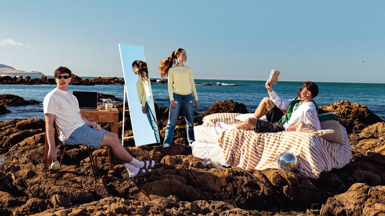 three students on rocks by the beach, with a bed, mirror and chair
