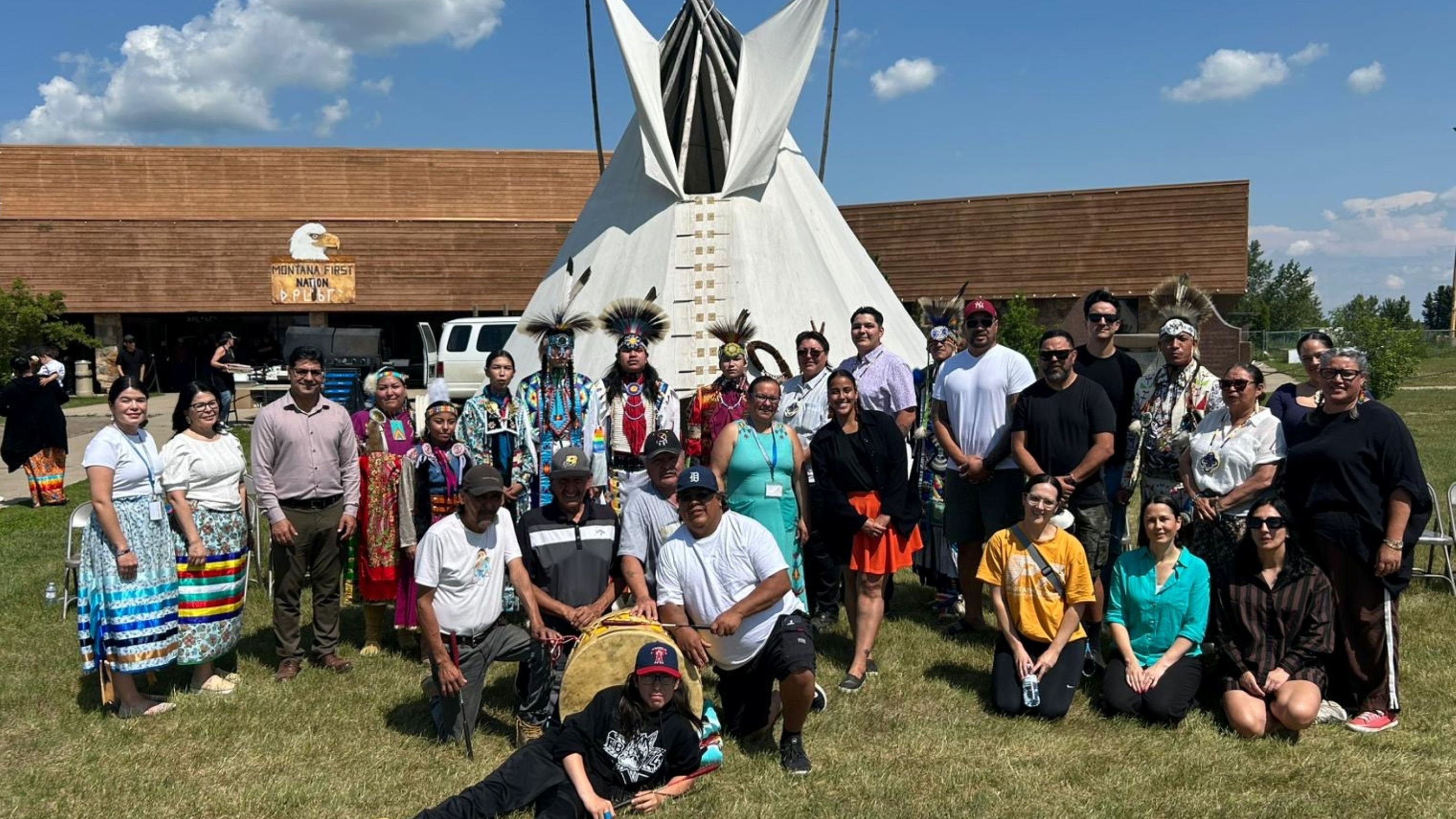 A group of people standing outside in front of a tall white tipi. Several individuals wear traditional Indigenous Canadian regalia.